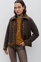 Chaqueta de lana merino hervida en color marrón, con diseño estructurado y cuello amplio. Presenta detalles de bordado artesanal en contraste color mostaza a lo largo del frente, cuello y puños, junto con botones forrados en la misma tela.