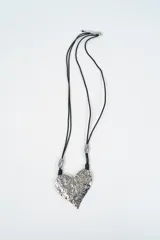Collar largo con cordón negro y dije de corazón grande con textura irregular en tono plateado, decorado con cuentas metálicas plateadas en los extremos del cordón.