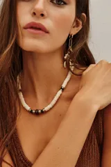 Collar de cuentas de concha de río en color crema, intercaladas con pequeñas piezas metálicas doradas, cuentas de vidrio verde y una cuenta central de vidrio en tono borravino.