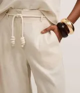 Pantalón largo de cintura alta color beige, confeccionado en tejido viscolino, con trabillas en la cintura, cinturón de cuerda para lazo, cierre tradicional y pliegues.