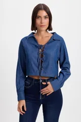 Camisa crop de denim azul con cuello camisero y cierre frontal mediante cordones ajustables entrelazados en ojales. Presenta mangas largas con puños rectos y corte entallado.