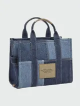 Cartera tipo tote bag Marc Jacobs modelo The Small Traveler Tote, confeccionada en denim con diseño de paneles en distintos tonos de azul. Cuenta con asas superiores, correa de hombro ajustable y extraíble, y el texto "THE TOTE BAG MARC JACOBS" estampado en la parte frontal.