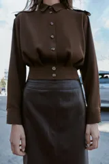 Camisa corta de cuello solapa y manga larga, con trabillas decorativas en los hombros. Presenta pliegues delanteros, elástico en la espalda y cierre frontal con botones.