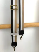 Correa ajustable para cartera confeccionada en cinta tejida con diseño de rayas en negro, blanco y beige, con punteras de cuero sintético negro y herrajes metálicos.