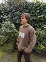 Sweater de tejido de punto color marrón, con cuello redondo y diseño holgado. Presenta un detalle bordado en el centro del pecho que representa la bandera de Uruguay.