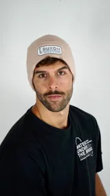 Gorro tipo beanie de punto acanalado en color beige, con parche rectangular frontal que incluye el logo de la marca Duxon.