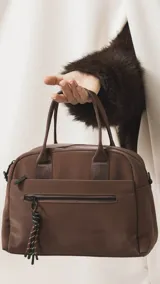 Cartera de mano color beige, confeccionada en tela impermeable, con doble asa corta y correa larga ajustable y desmontable. Presenta cierre principal, bolsillo frontal con cierre y detalle de colgante decorativo.