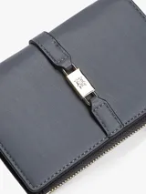 Billetera bifold Tommy Hilfiger de diseño compacto en color gris. Presenta un cierre superior con cremallera y un detalle frontal decorativo con broche metálico que incluye el logo de la marca. Cuenta con múltiples compartimentos internos para tarjetas y billetes.