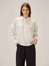 Camisa de lino de corte holgado, color blanco, con cuello clásico, cierre frontal de botones y detalle de pespuntes en zigzag en los hombros y puños.