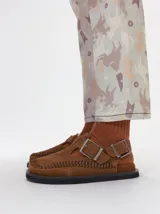 Slipper tipo guillermina confeccionada en gamuza color marrón, con plataforma de goma negra y doble hebilla ajustable. Presenta un pespunte decorativo en el borde inferior y cuenta con interior recubierto en cuero.