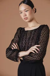 Blusa de encaje negro con diseño de flores, cuello redondo y mangas largas con terminación en puño con detalle de pestañas. Presenta una silueta ajustada y femenina.