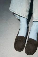 Mocasín de serraje color marrón oscuro, con detalle de fruncido y lazo en la parte delantera, y pasadores decorativos en los laterales.