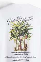 Remera de algodón blanca con cuello redondo y hombros caídos. Presenta un pequeño estampado tipográfico en el pecho con la frase 'Puerto Vallarta Beach Bar'.