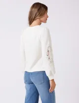 Buzo blanco de modal, corte clásico, con mangas largas abullonadas y apliques de crochet floral en los hombros y parte superior de las mangas.