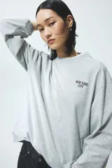 Top oversized de punto de algodón suave en color gris melange, con diseño estampado de letras pequeñas en el pecho. Presenta manga larga, cuello redondo ribeteado y hombros caídos.