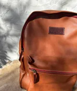 Mochila de cuero negro, con forro interior de lino y herrajes de bronce. Presenta un bolsillo frontal con cierre y un parche de cuero marrón con logo grabado.