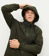 Campera rompevientos de color gris oscuro, con capucha ajustable mediante cordones, cierre frontal por cremallera y bolsillos laterales con cierre. Presenta puños elásticos y un diseño liso y minimalista.