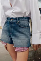 Shorts de jean tiro medio, corte mom fit, con bajo deshilachado y forro de bolsillos delanteros estampado a cuadros que asoma por debajo.