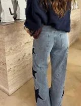 Jean de tiro alto con pierna amplia de corte recto, confeccionado en denim de lavado claro. Presenta aplicaciones de estrellas en contraste con bordes decorados con tachas metálicas.