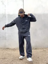 Conjunto de dos piezas compuesto por un buzo con capucha y cierre frontal y un pantalón de jogging, ambos confeccionados en tejido con efecto acid wash en color gris oscuro. El buzo cuenta con bolsillos frontales y logo bordado, mientras que el pantalón presenta un corte holgado y logo bordado en la pierna.