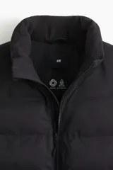 Chaleco acolchado de color negro, confeccionado en tejido guateado repelente al agua y rompevientos. Presenta un diseño con cuello elevado, cierre frontal completo con protección para el mentón y bolsillos laterales con cierre. Posee un corte estándar y está forrado internamente.
