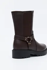 Bota tipo biker de caña media en cuero sintético negro, con panel elástico lateral, suela gruesa dentada y arnés decorativo con herraje metálico.