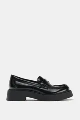 Mocasín de cuero negro con suela gruesa tipo plataforma, detalle de antifaz con aplique metálico dorado y costuras reforzadas en la puntera.