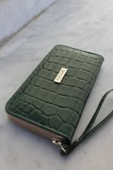 Billetera tipo sobre con correa de muñeca, confeccionada en cuero con textura de reptil en color marrón. Cuenta con cierre perimetral y placa metálica con el logo de la marca en el frente.