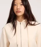 Saco color crema, de corte slim, confeccionado en algodón con viscosa certificada, con cuello con capucha y cordón ajustable, cierre frontal con cremallera y forro de fleece.