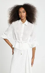 Camisa de rayas verticales blanca de Le Lis Blanc, modelo Regina, confeccionada en tejido fluido con ligera transparencia. Presenta corte recto, cuello clásico, mangas largas con volumen y puños ajustados, cierre frontal de botones y detalle fruncido con cordón en la cintura.