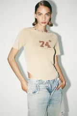Camiseta color beige de algodón con efecto lavado, corte regular, cuello redondo con tachas metálicas, mangas cortas y estampado frontal con el número 74 y una estrella.
