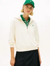 Sweater de felpa color blanco marfil, con cuello alto y cierre frontal de media cremallera. Presenta mangas largas con puños y dobladillo acanalados, y un pequeño logo script bordado en verde en el pecho.