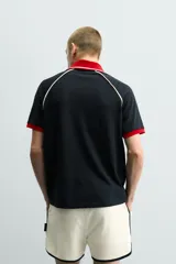 Polo de corte relajado confeccionado en tejido de algodón con estructura abierta. Presenta cuello polo con abertura frontal en V, mangas cortas tipo ranglán y acabados en rib a contraste en color rojo y blanco.