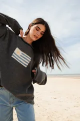 Sweater de hilo de algodón color verde militar, con cuello redondo y manga larga. Presenta un diseño tejido de la bandera uruguaya en el frente.