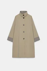 Trench de corte oversize color beige, con cuello alto, cierre frontal de botones y mangas largas con puños vueltos estampados a cuadros. Cuenta con bolsillos delanteros de vivo.