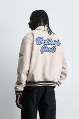 Cazadora estilo varsity de tejido efecto ante en color beige. Presenta cuello solapa, cierre frontal con botones a presión y puños y ruedo con acabado en rib a rayas negras. Incluye parches bordados decorativos en el pecho y la manga, además de bolsillos laterales de doble vivo.
