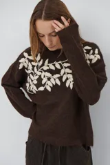 Buzo de tejido de punto en color beige, con cuello redondo y mangas largas. Presenta un delicado bordado de ramas y hojas en color crema en la parte superior del frente.