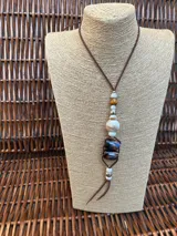 Colgante largo con cordón de cuero marrón y diseño central compuesto por diversas cuentas de piedra, cerámica y metal en tonos tierra, blanco y azul oscuro.
