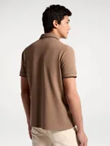 Chomba de piqué color tostado con cuello y puños en contraste negro. Presenta cierre frontal con botones y un pequeño bordado en el pecho.