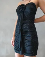 Vestido corto strapless negro con diseño drapeado en toda la prenda, detalle de aplique floral en el centro del escote y caída en cascada con volados frontales. Presenta pequeños puntos brillantes distribuidos por toda la superficie.