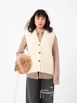 Chaleco largo color crema, tejido en punto, con cuello en V, cierre frontal con botones marrones a contraste y bolsillos de parche.