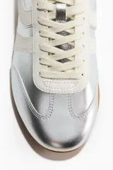 Championes deportivos de estilo urbano, color plateado con bandas laterales y cordones en tono crudo, y suela de goma marrón.