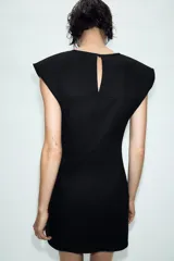 Vestido corto de estilo militar en color negro, con cuello redondo, hombreras y manga sisa. Presenta un diseño frontal con falsos botones dorados dispuestos en filas horizontales. Cierre lateral con cremallera oculta y abertura en la espalda con botón.