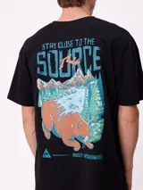 Remera de corte oversize color crema con estampado gráfico en la espalda que muestra un oso, un paisaje natural y el texto 'Stay close to the source'.