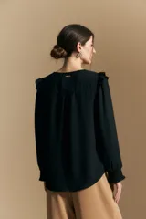 Camisa negra de manga larga con escote en V y cierre frontal de botones. Presenta detalles de volados en los hombros, frunces en la espalda y puños elastizados que le otorgan una silueta relajada y fluida.