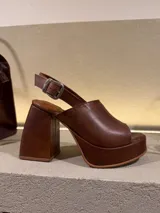 Sandalia de cuero marrón oscuro con plataforma y taco ancho y alto. Presenta una capellada abierta en la punta y una tira ajustable con hebilla metálica en el tobillo.