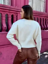 Suéter de punto color crema, con cuello redondo, manga larga y cierre frontal parcial que deja ver parte del abdomen.