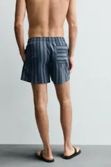 Bañador de hombre de largo medio, color azul claro con rayas verticales en azul oscuro. Posee cintura elástica ajustable con cordón, bolsillos laterales y un bolsillo trasero de plastrón.