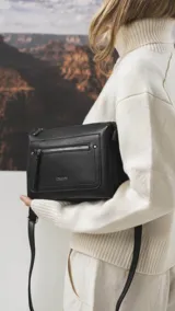 Bandolera de cuero sintético negro con textura granulada, cuenta con un bolsillo frontal con cierre, correa ajustable y desmontable, y herrajes metálicos.