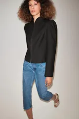 Chaqueta negra de cuello redondo, manga larga con hombreras abullonadas y cierre frontal con ganchos metálicos.